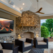 A large TV is hung on an outside brick exterior next to a fireplace and patio seating.