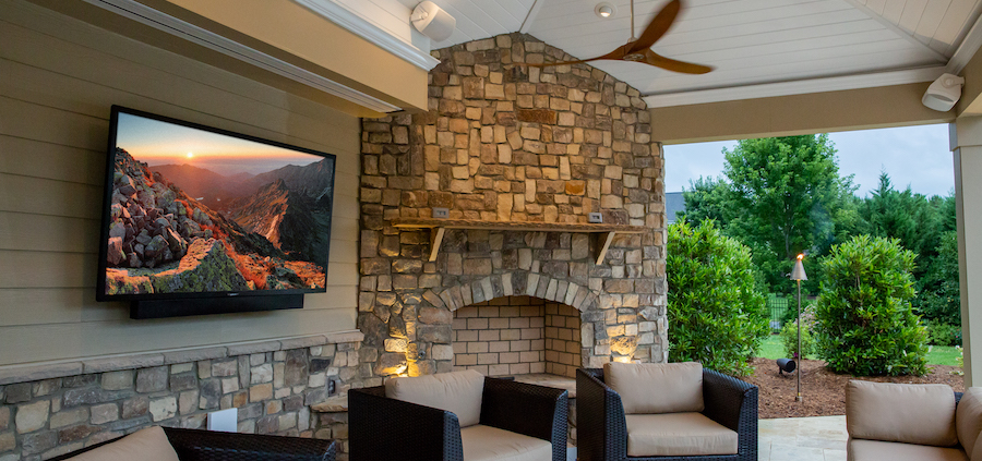 A large TV is hung on an outside brick exterior next to a fireplace and patio seating.