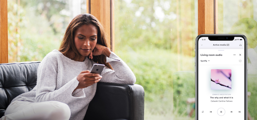 A woman relaxes on a couch, looking at her phone. A side image shows the Crestron Home app with Spotify playing audio in the living room.